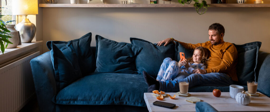 A father and daughter sit together on a sofa, both engaged with a smartphone, sharing a moment of connection.